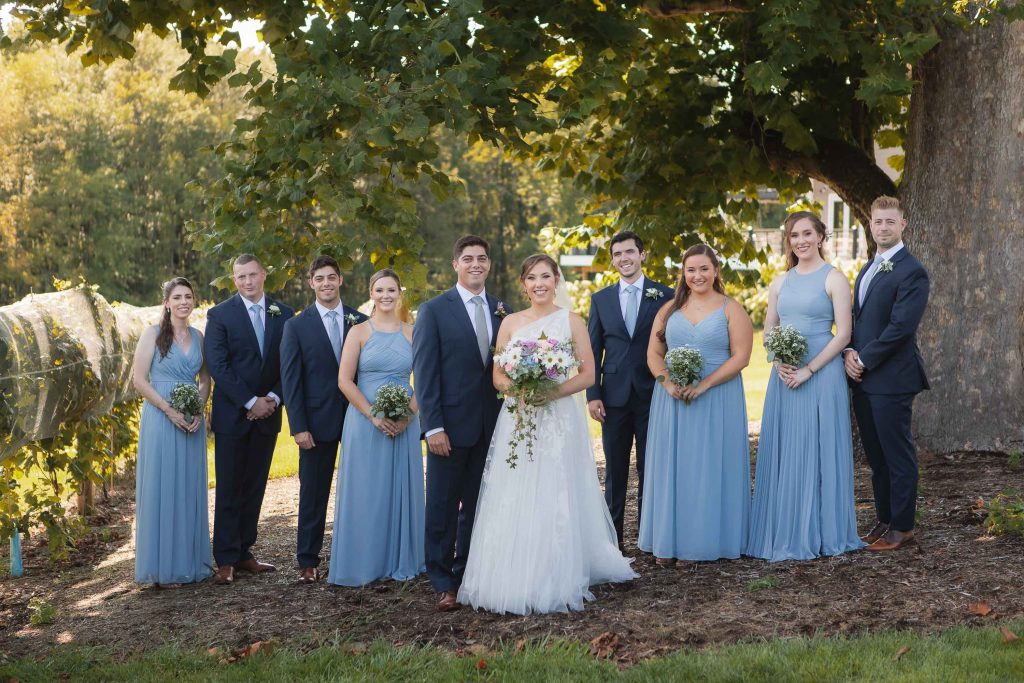 A group of bridesmaids and groomsmen posing in front of a tree at Fleetwood Farm Winery for their wedding portrait.
