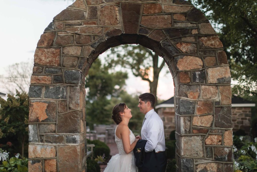 A bride and groom share a tender moment in front of a stone archway at Fleetwood Farm Winery during their portrait session on their wedding day.