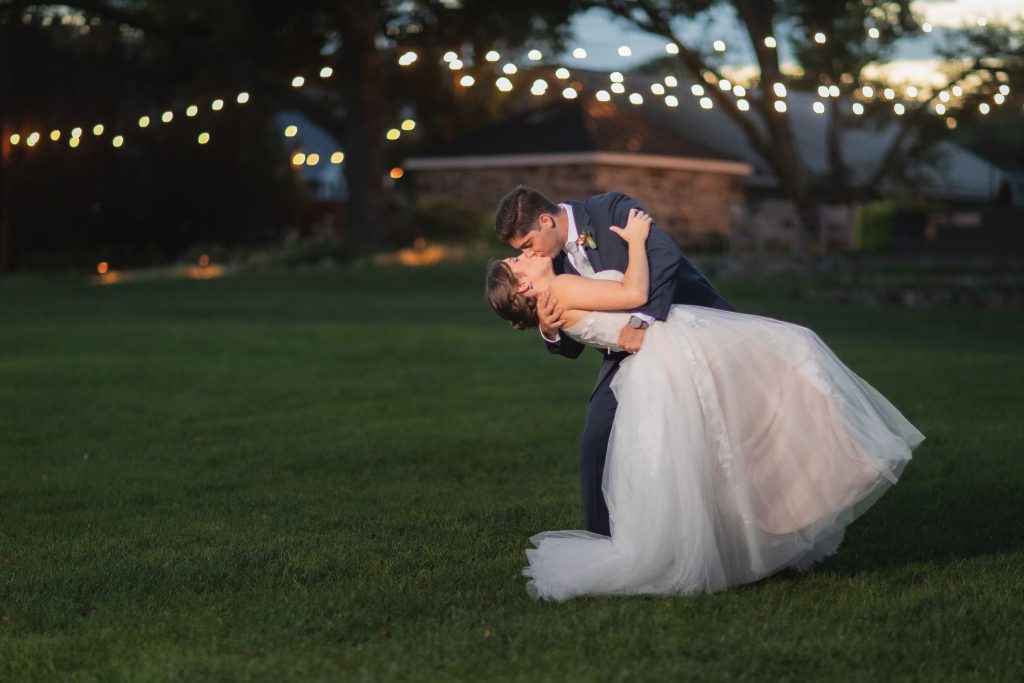 A bride and groom sharing a romantic portrait at Fleetwood Farm Winery during their wedding, as they passionately kiss under the soft glow of string lights in the lush grass.