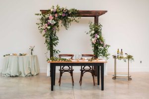 The Pink Poppy event vendor offers a table set up with flowers and a wooden arch.