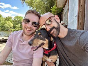 Two men with sunglasses and a dog on a dock.