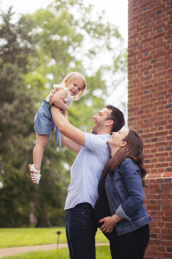 A family is holding their baby in front of a brick building for a portrait session.