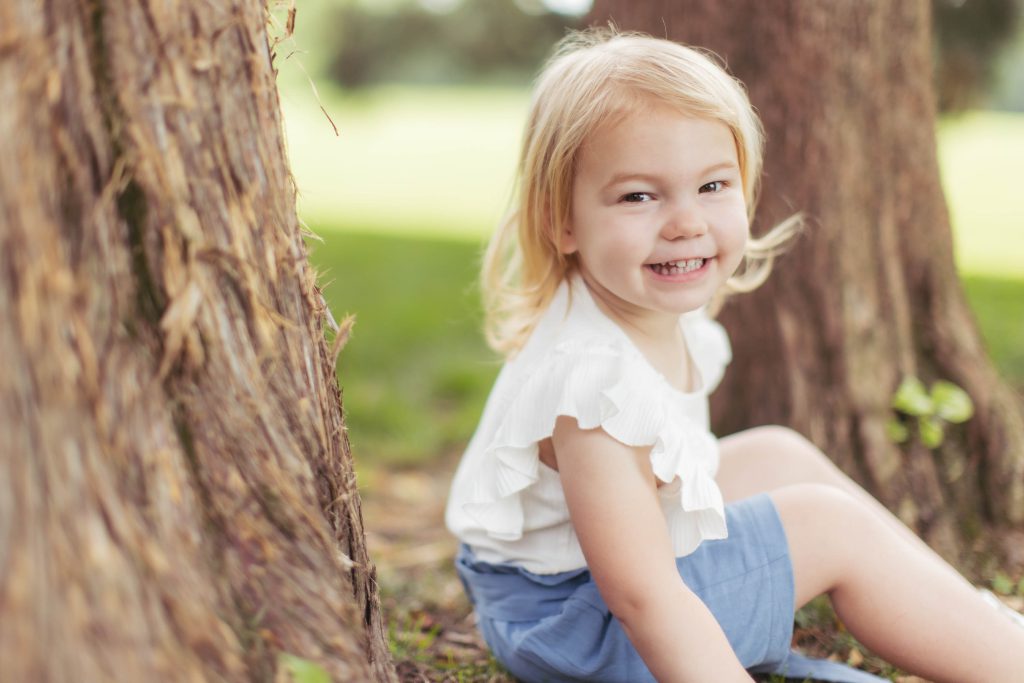 A little girl sitting in the grass next to a tree for family portraits.