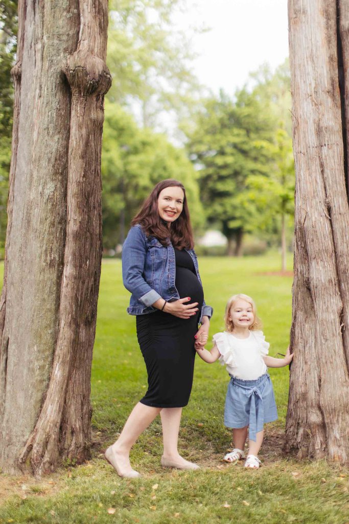A pregnant woman and her daughter pose for family portraits in front of a tree.