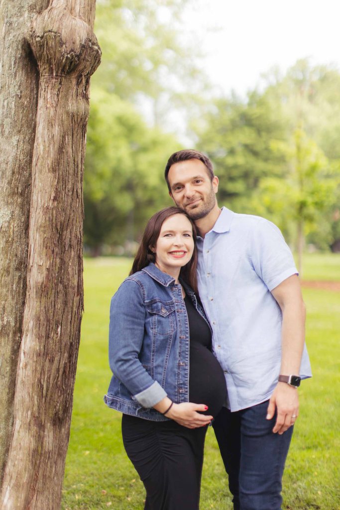 A pregnant couple standing next to a tree in a park, capturing family portraits.