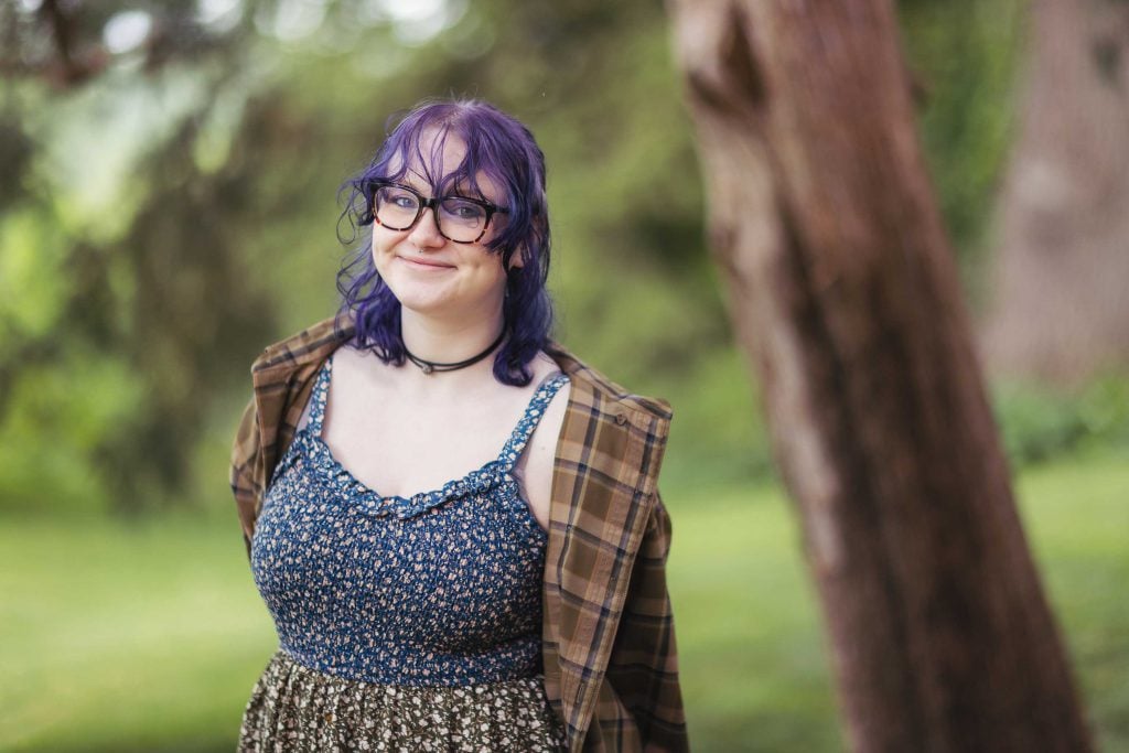 A high school senior woman with purple hair wearing glasses and a plaid dress.