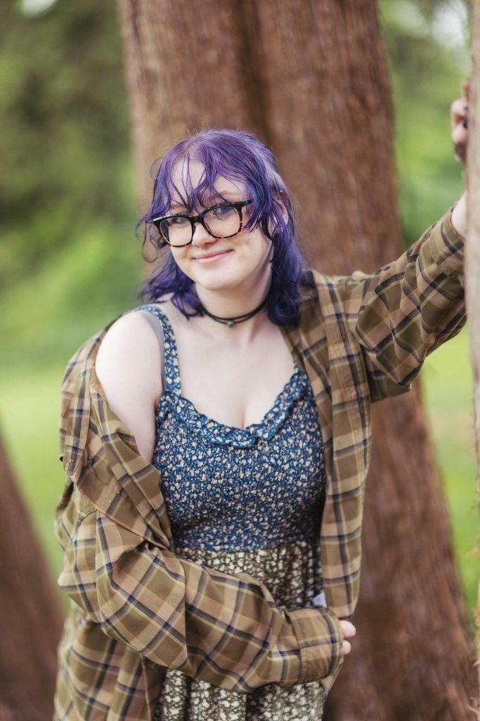 A high school senior with purple hair leaning against a tree.