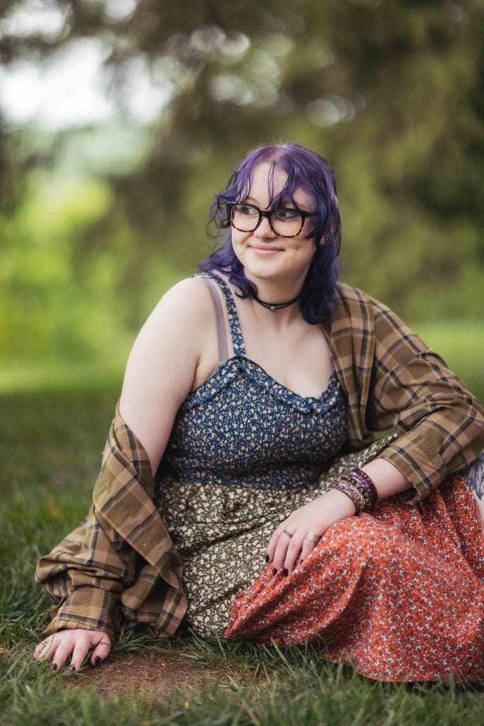 A high school senior wearing glasses and a plaid dress sitting on the grass during graduation at a Bel Air mansion.