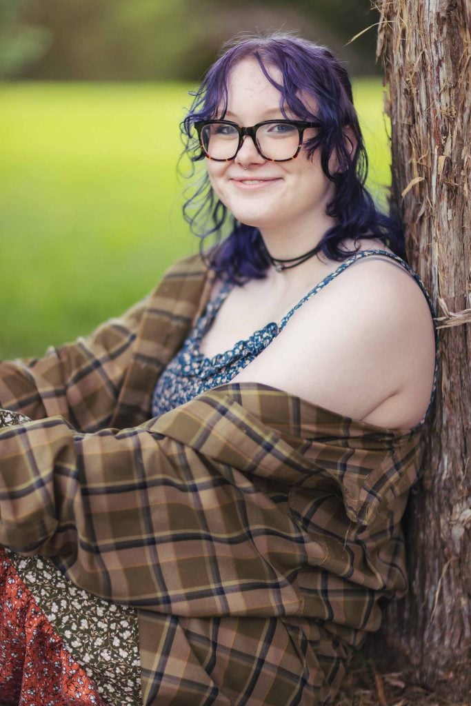 A woman wearing glasses and a plaid shirt posing for a portrait leaning against a tree.