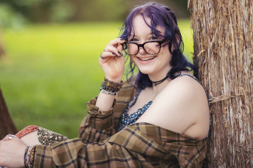 A woman wearing glasses leaning against a tree, in a graduation portrait.