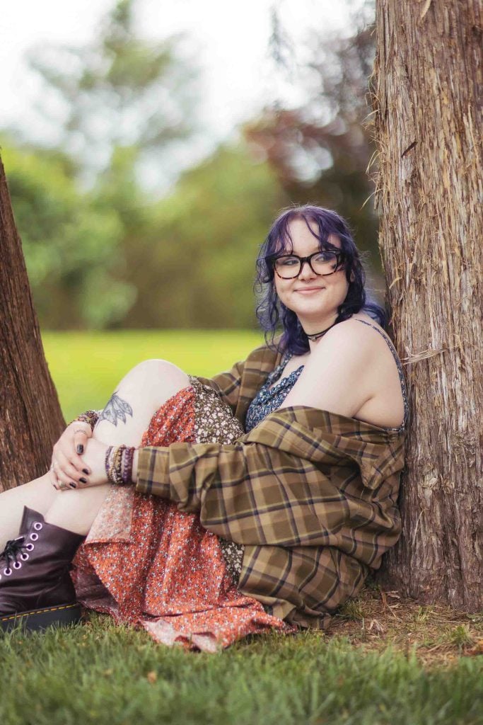 A High School Senior sitting next to a tree wearing glasses and a plaid skirt, preparing for graduation at the Bel Air Mansion.