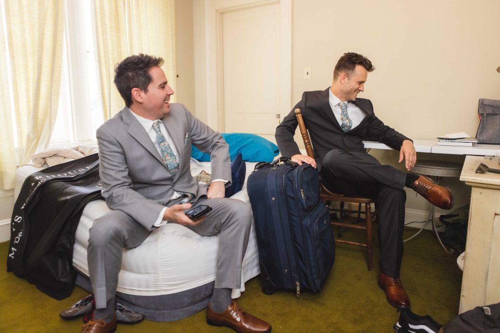 Two men in suits candidly preparing for a wedding on a bed in a room.