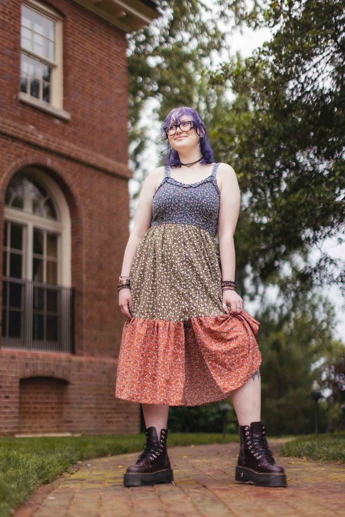 A High School Senior with purple hair standing in front of a brick house.
