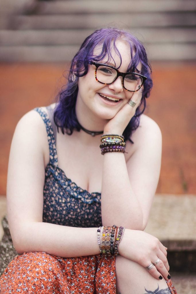 A high school senior with purple hair and glasses sitting on steps.
