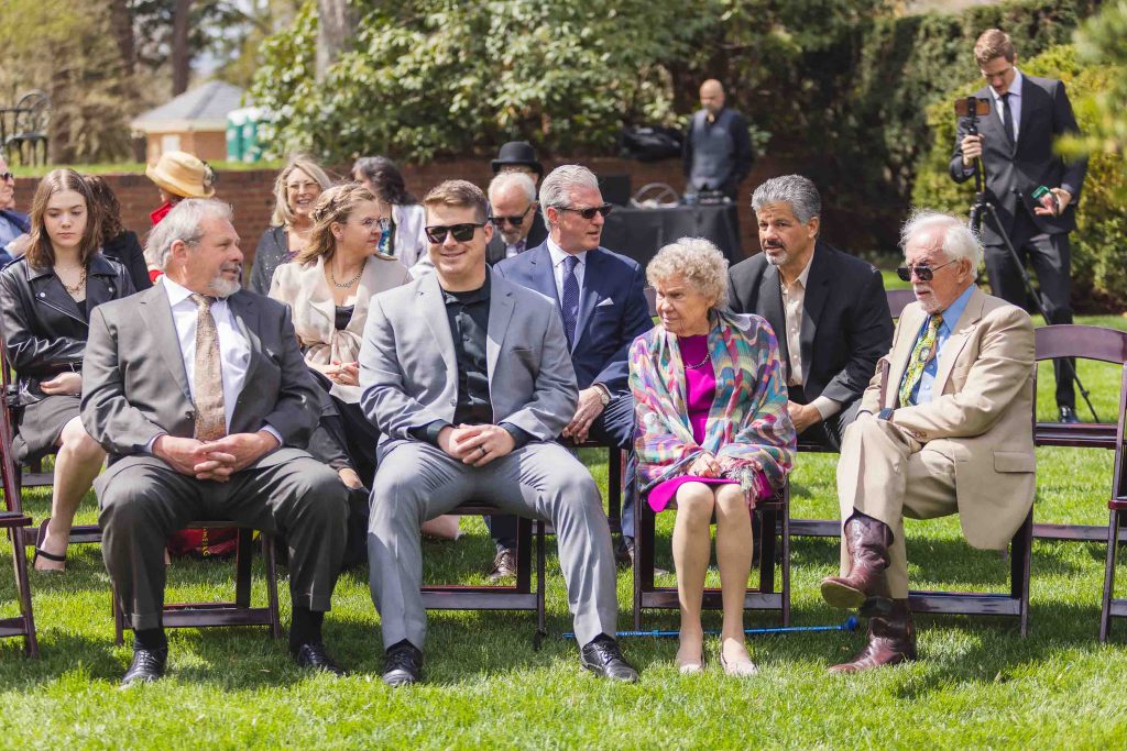 A group of people sitting on chairs during a wedding ceremony in a grassy area.