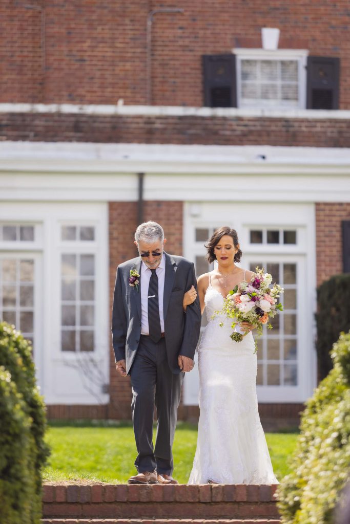 A bride and her father walking down the steps of a brick house during the wedding ceremony.