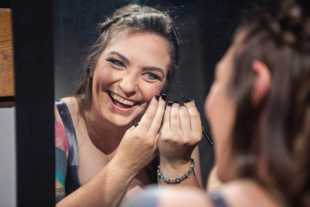 A woman smiling candidly in front of a mirror during wedding preparations.