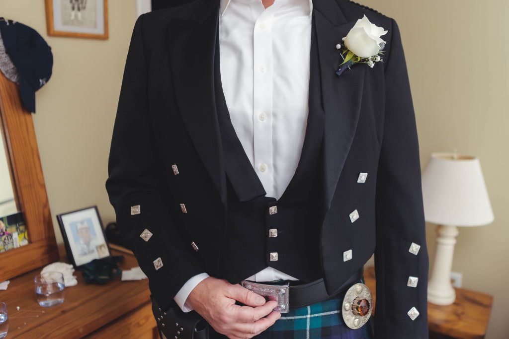 A groom in a kilt is preparing for his wedding, standing candidly in front of a mirror.