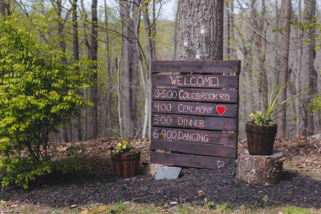 A wooden sign displaying wedding details placed next to a tree in the woods.