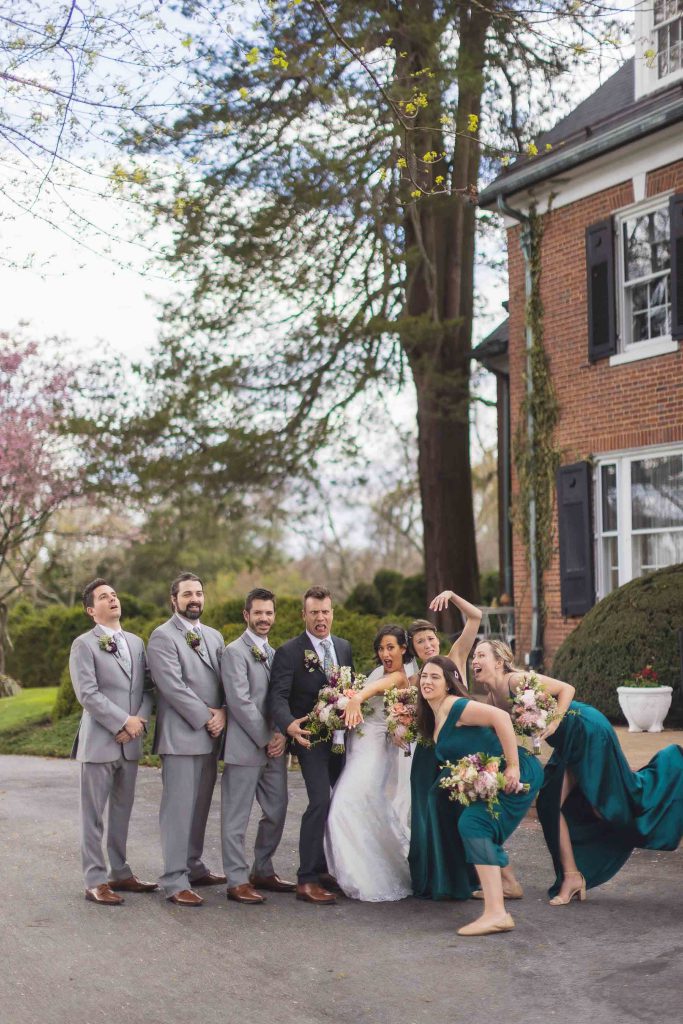 A group of bridesmaids and groomsmen pose for a wedding portrait in front of a house.
