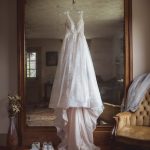 A detailed wedding dress hangs in front of a mirror in a room.