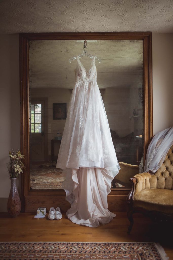 A detailed wedding dress hangs in front of a mirror in a room.