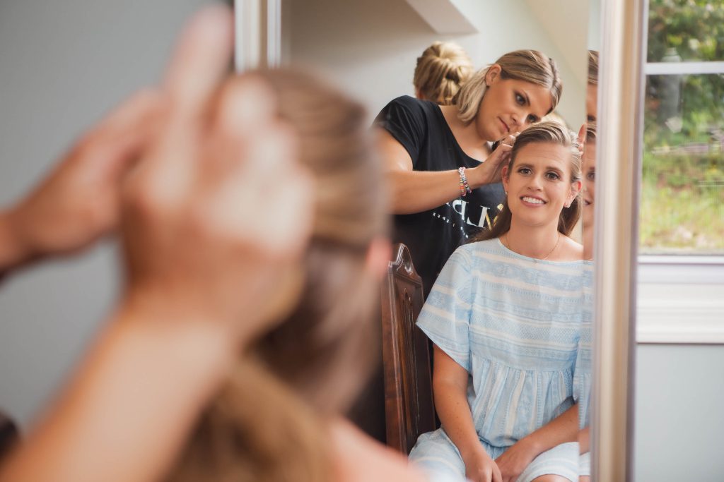 A woman candidly preparing for her wedding, getting her hair done in front of a mirror.