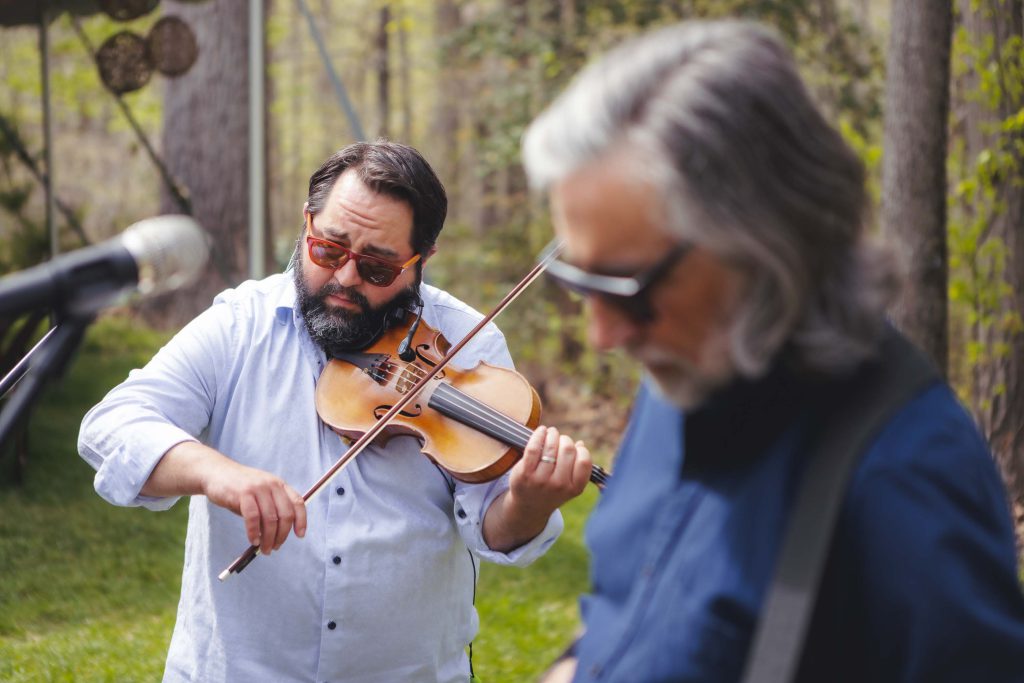 Two bearded men playing violin in a wooded area during a candid moment.