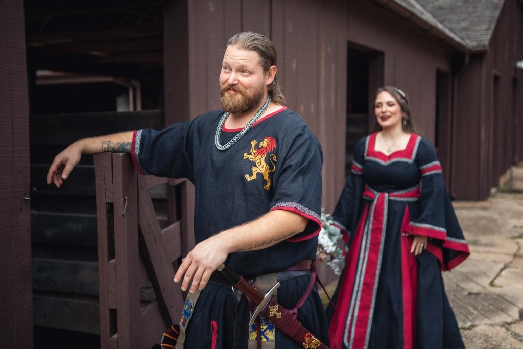 A candid man and woman dressed in medieval clothing are standing in front of a barn in preparation for their wedding.