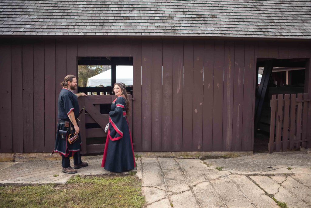 A candid couple dressed in medieval clothing prepares for their wedding in front of a barn.