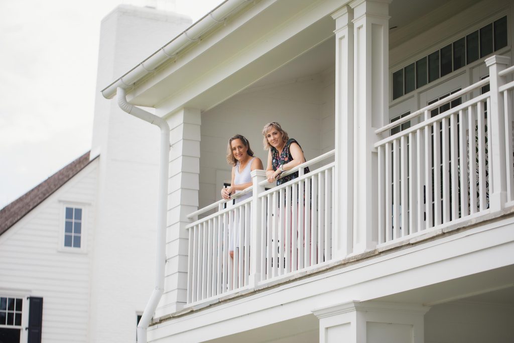 Two women candidly preparing for a wedding on the balcony of a white house.