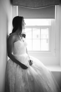 A candid bride in her wedding dress sits by a window, preparing for her big day.