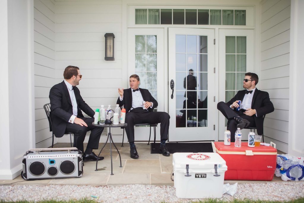 Three men in tuxedos candidly preparing for a wedding on the porch of a house.