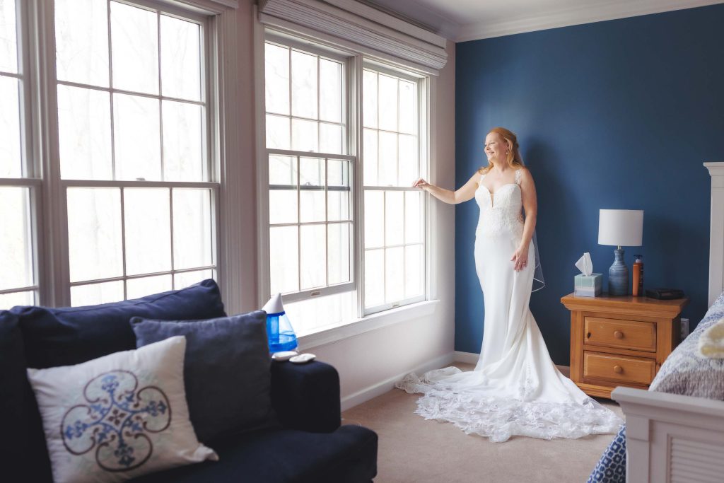 A candid bride prepares for her wedding in front of a window in her bedroom.