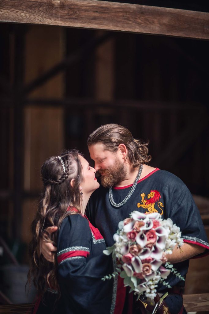 A medieval groom and bride share a passionate kiss during their wedding portrait session in front of a wooden barn.