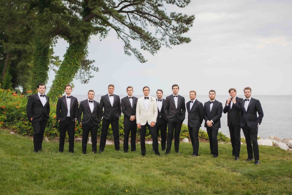A group of groomsmen in tuxedos standing in front of the ocean for a wedding portrait.