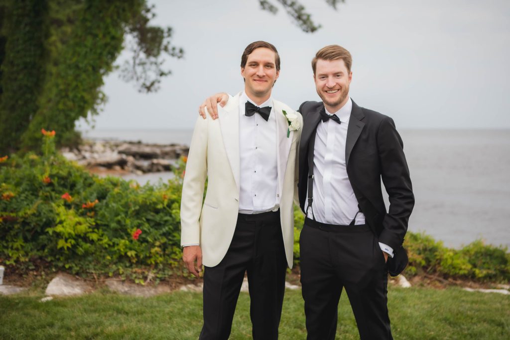 Two men in tuxedos posing for a wedding portrait next to the water.