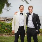 Two men in tuxedos posing for a wedding portrait next to the water.