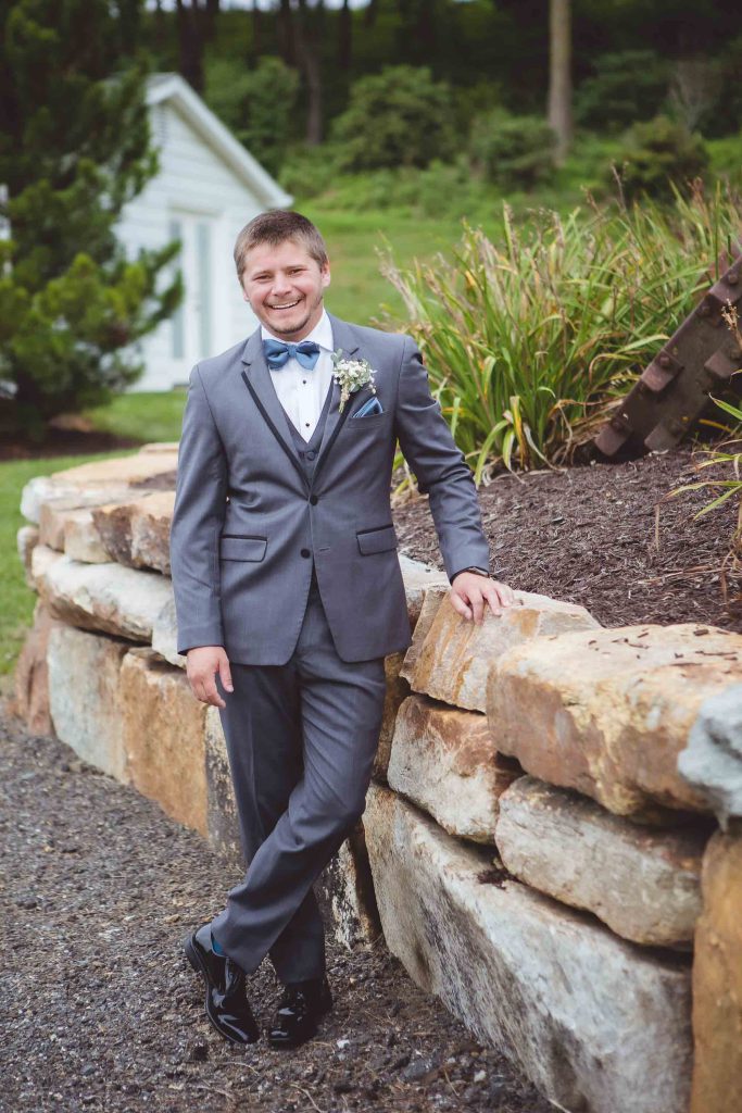 A groom in a wedding gray suit leaning against a stone wall.
