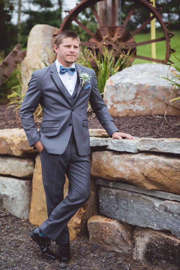 A groom in a gray suit posing for a wedding portrait against a stone wall.