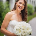 A bride smiles while holding a bouquet of white flowers on her wedding day.