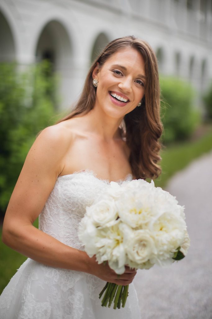 A bride smiles while holding a bouquet of white flowers on her wedding day.