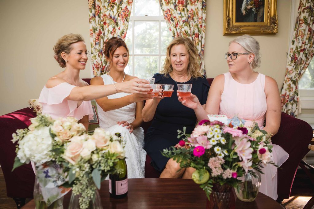 A group of bridesmaids in candid celebration, toasting with wine glasses during wedding preparation.