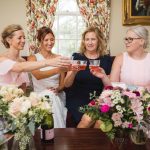 A group of bridesmaids in candid celebration, toasting with wine glasses during wedding preparation.