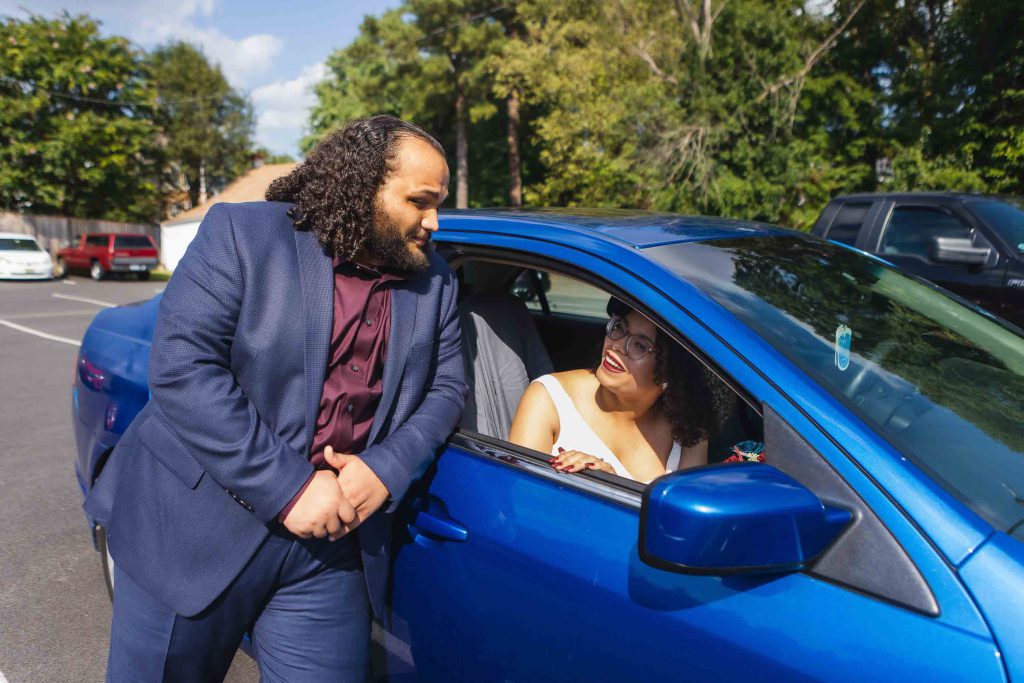 A man and a woman standing next to a blue car during a wedding preparation.