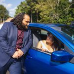 A man and a woman standing next to a blue car during a wedding preparation.