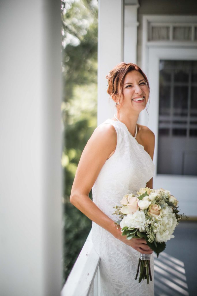 A wedding bride smiles while holding her bouquet on the porch.