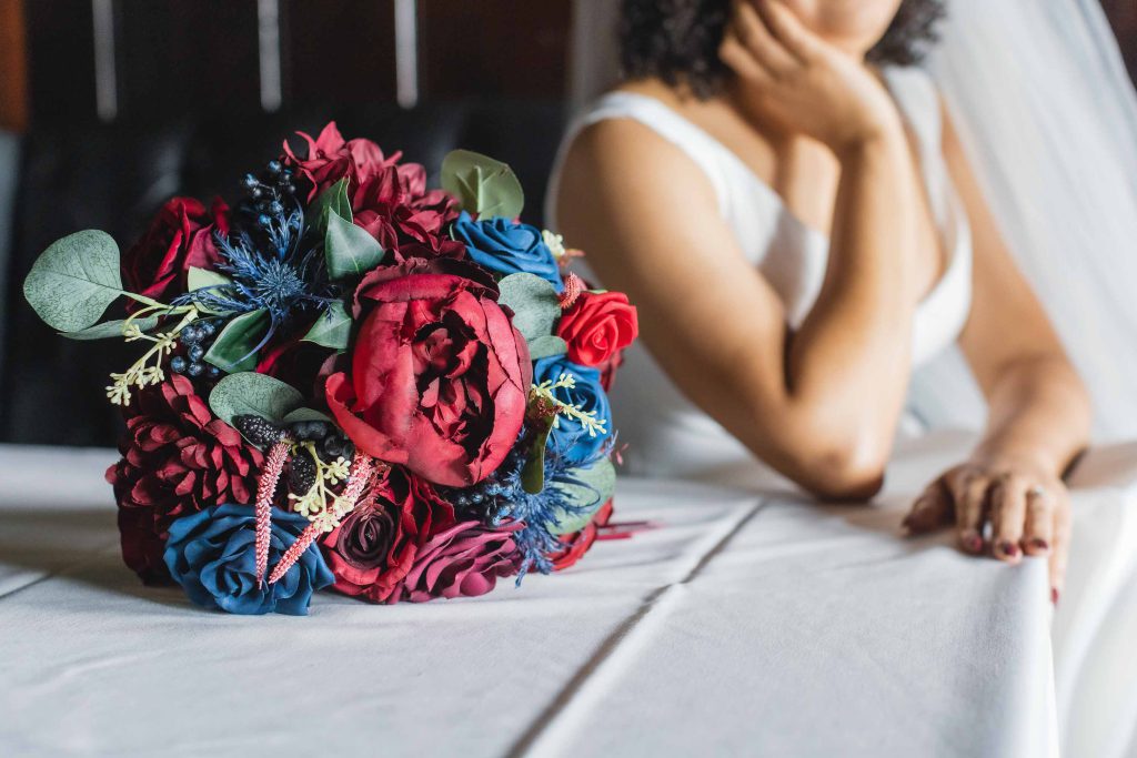 A bride sits on a table with a bouquet of red and blue flowers, highlighting wedding details.