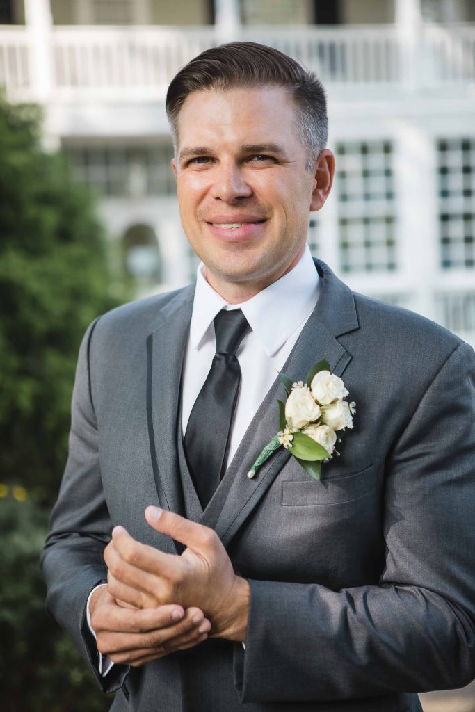 A groom in a gray suit smiles in front of a house, capturing a wedding portrait.