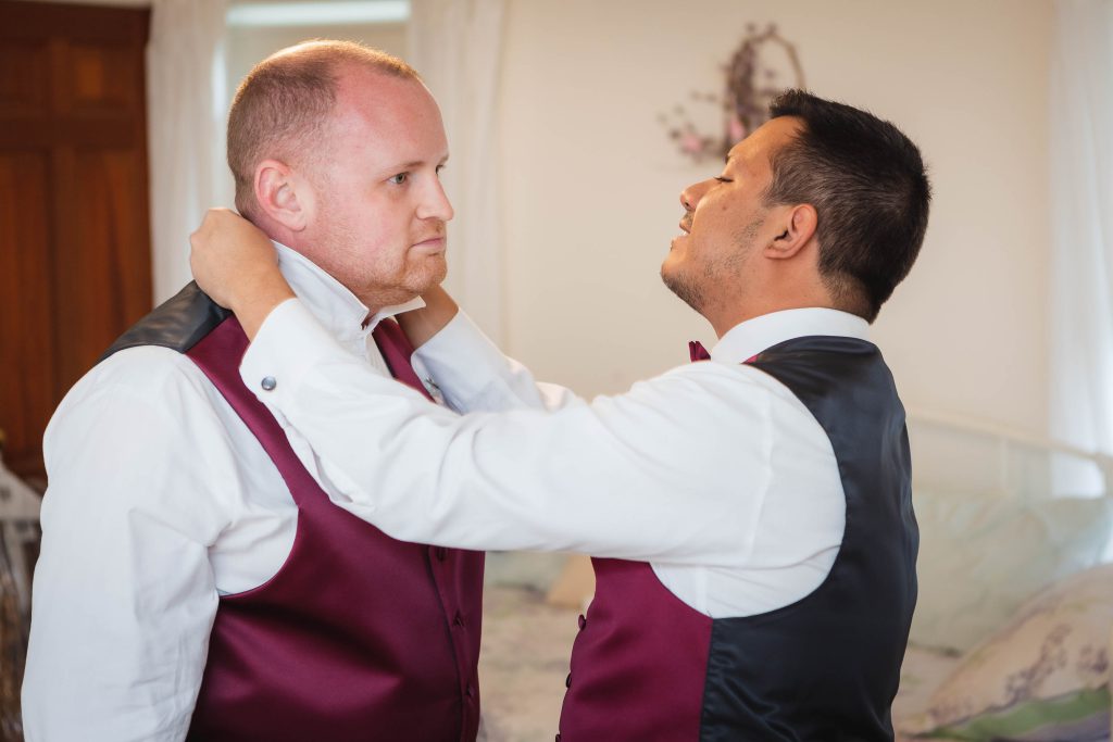 Two men preparing for a wedding while candidly adjusting each other's vests in a bedroom.